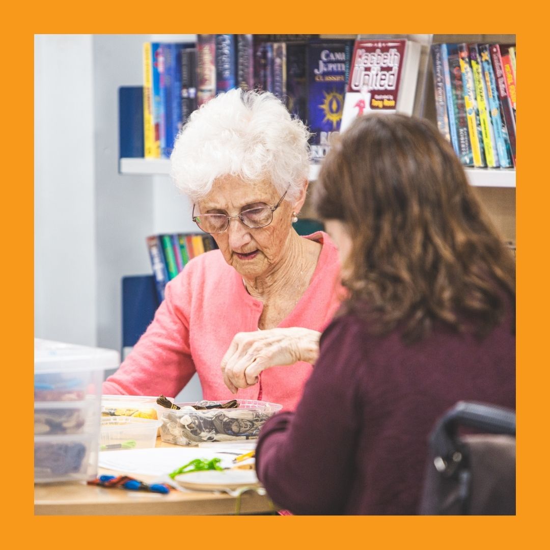 A woman with white hair and a woman in a wheelchair work on a craft at a table in the library