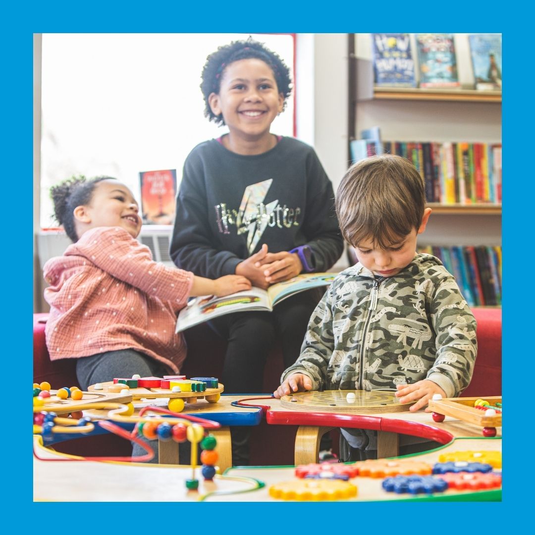 A child and two pre-schoolers enjoy books and table manipulatives in a library