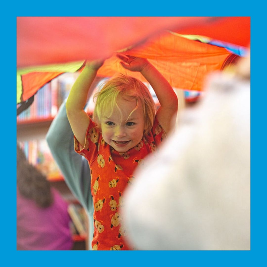 A photo of a smiling child raising its hands while standing under a colourful parachute