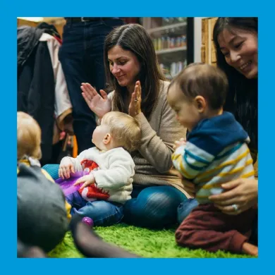 A photo of toddlers sitting on the floor in parents' laps, clapping and smiling